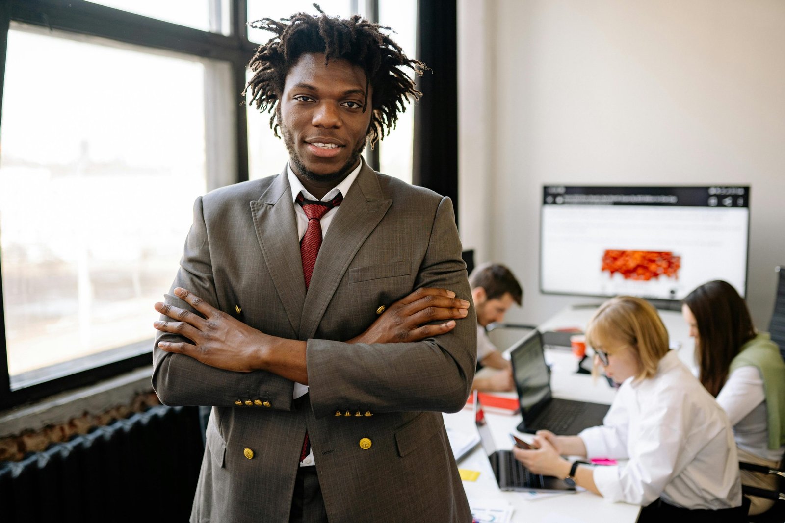 Confident businessman with arms crossed in a bright office environment with colleagues working in the background.